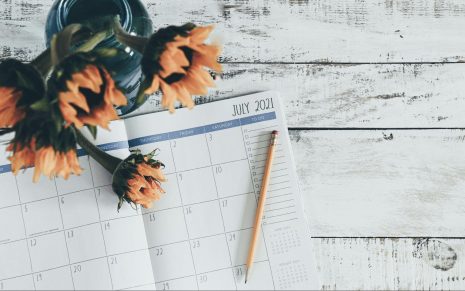 Planner on a desk open to July with a pencil and a vase of flowers nearby